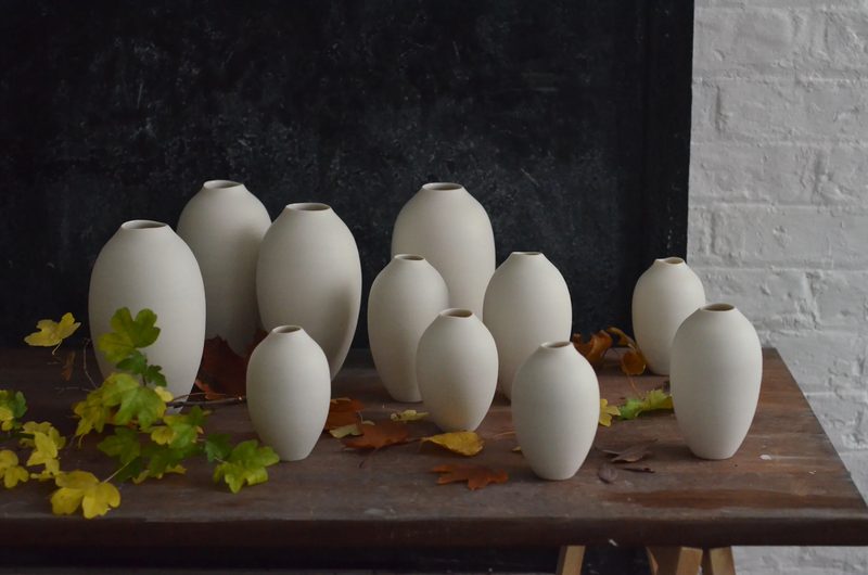 A cluster of small hand-thrown porcelain bud vases with autumn leaves on a wooden table